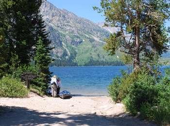 Visitor launching a blue kayak at the rocky Jenny Lake Boat Launch with conifer trees on either side of the ramp area.