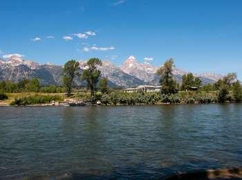 A landscape photo with the snake river in the foreground, ferry boat in the river on the opposite bank, and the whitewashed buildings of Menors Ferry with the Teton Range towering above.