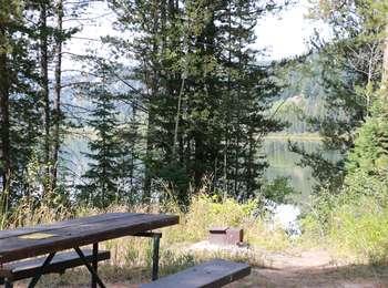 Picnic table at Two Ocean Lake Trailhead with fire grate below and view through lodgepole pines onto Two Ocean Lake