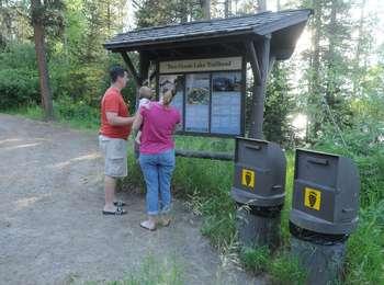 Two visitors with a baby at the Two Ocean Lake trailhead sign shaded by lodgepole pines.