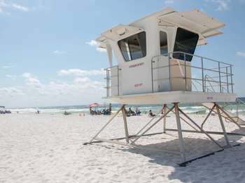 A white structure on the white sand beach. In the background, people walk and lay on the sand, and play in turquoise water against a blue sky.