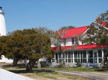 The distinctive red-tin roof of the historic Double Keepers' Quarters is nestled in with trees next to the Ocracoke Lighthouse.