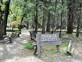 A grey wooden sign that reads Yosemite Cemetery is located next to a dirt path that is lined by zig-zagging wooden fences, and leads into the cemetery.