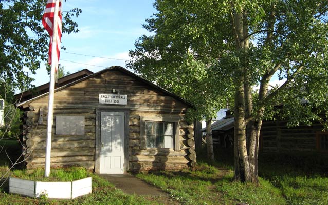 Eagle City Hall, a log cabin with a white door, an American flag post, and surrounded by trees. Eagle City Hall, a log cabin with a white door, an American flag post, and surrounded by trees.