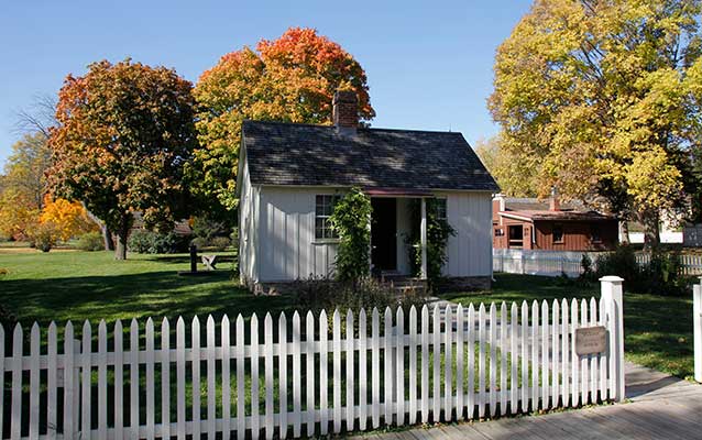 Trees in autumn foliage and a white picket fence surround a white cottage. Trees in autumn foliage and a white picket fence surround a white cottage.