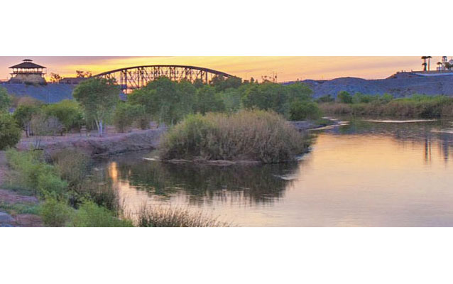 A tree-lined river, bridge in the distance, and the backdrop of a pastel evening sky. 