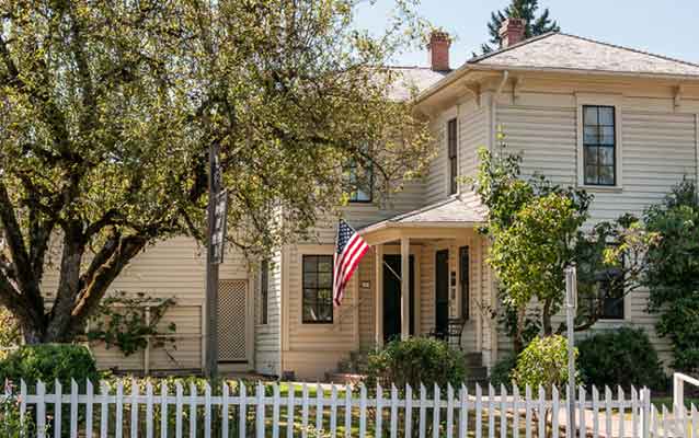 A United States flag flies from the front porch of an L shaped white two-story house.