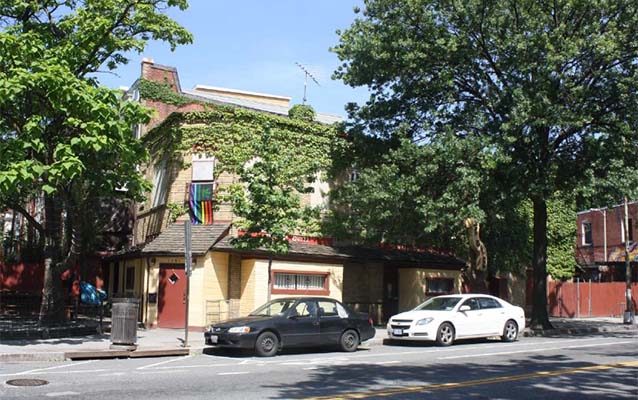 Exterior of the building; yellow brick with brick red door and a pride flag. 