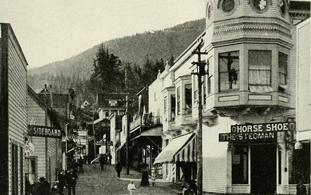 Black and white photo of a street lined with  shops with mountains in background. 