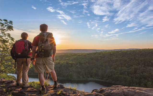 2 people stand on a cliff's edge, the sunset colors the valley below. 