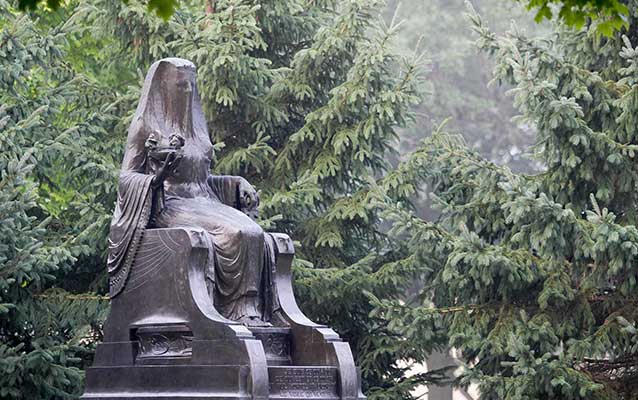 A cast bronze figure of a veiled goddess sits on a throne mounted on a concrete pedestal. A cast bronze figure of a veiled goddess sits on a throne mounted on a concrete pedestal.