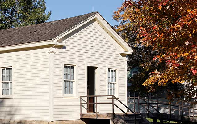 A one-story wooden building painted off-white has two windows and a central doorway. A one-story wooden building painted off-white has two windows and a central doorway.