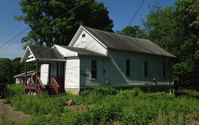 a modest one-story front-gable building with a concrete foundation, wood clapboard walls