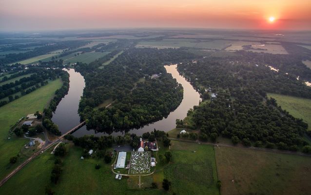 Sun setting upon meandering river surrounded by trees and clear cut plots.