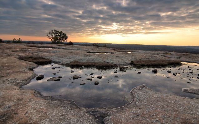 Sun setting on granite mountain with tree silhouette in the background and puddle in the foreground.