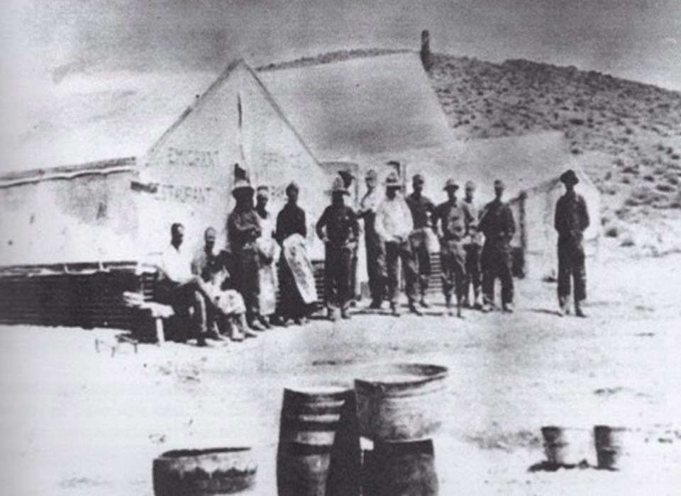 Black and white photo of men in front of tents with a desert hill behind and barrels in the foreground.