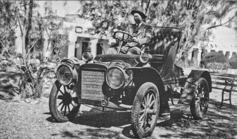 Man in old car with trees and a building