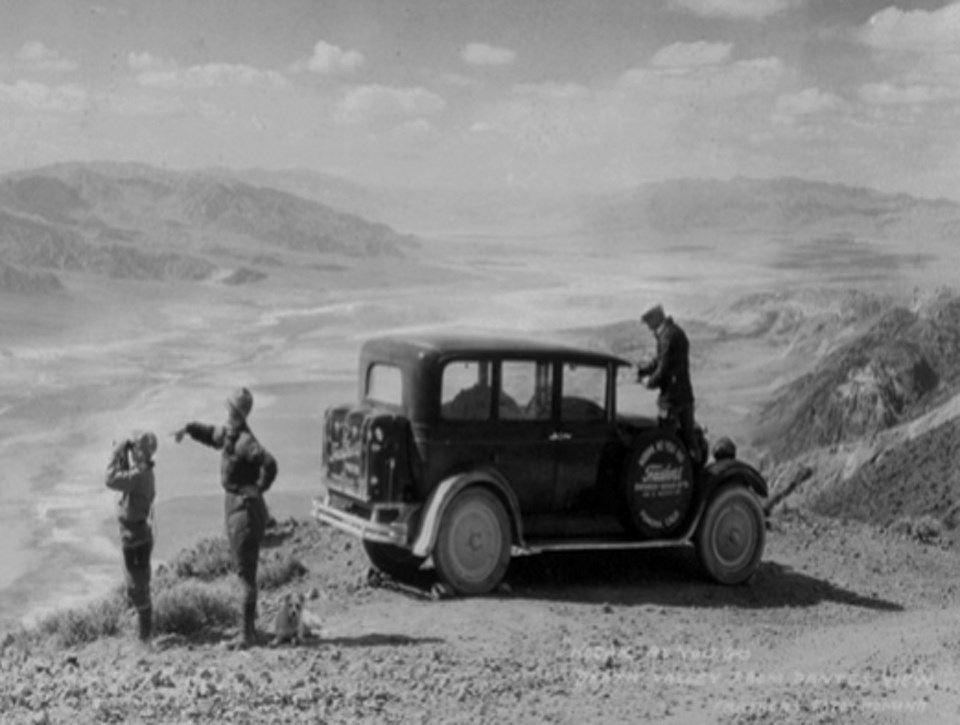 Three people next to old car with desert valley in background.