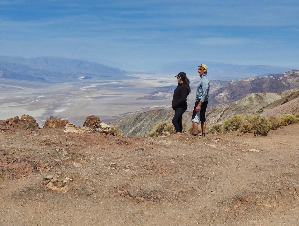 Three people next to old car with desert valley in background.