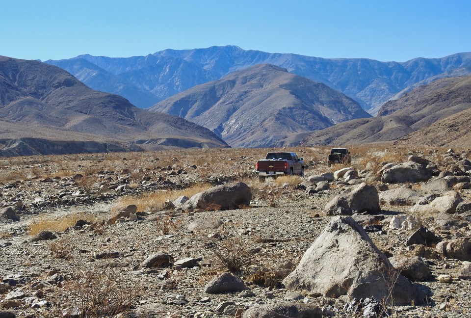 An old car drives on a dirt road with snow-capped peaks in background.