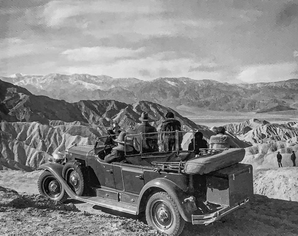 Old car with people parked overlooking desert landscape.