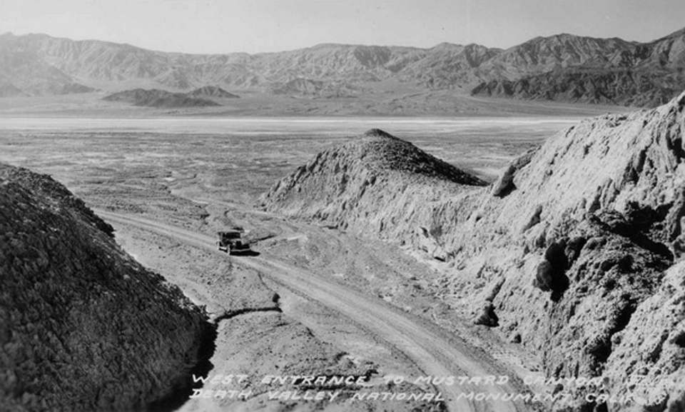 An old car drives on dirt road into dried mud canyon.