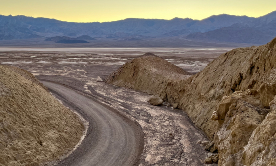 An old car drives on dirt road into dried mud canyon.