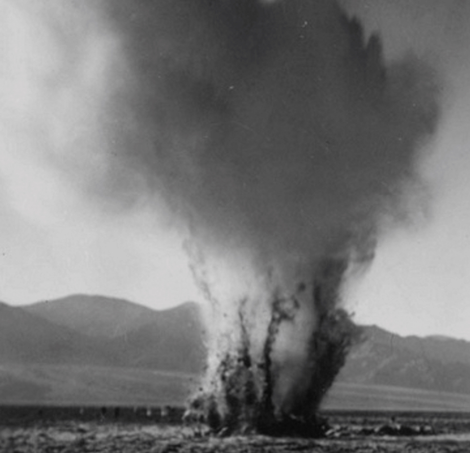 A blast of water and rock rises from flatland with mountains in background.