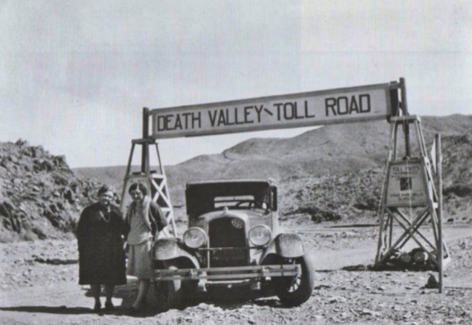 Two women stand next to old car in front of sign with hills in  background.