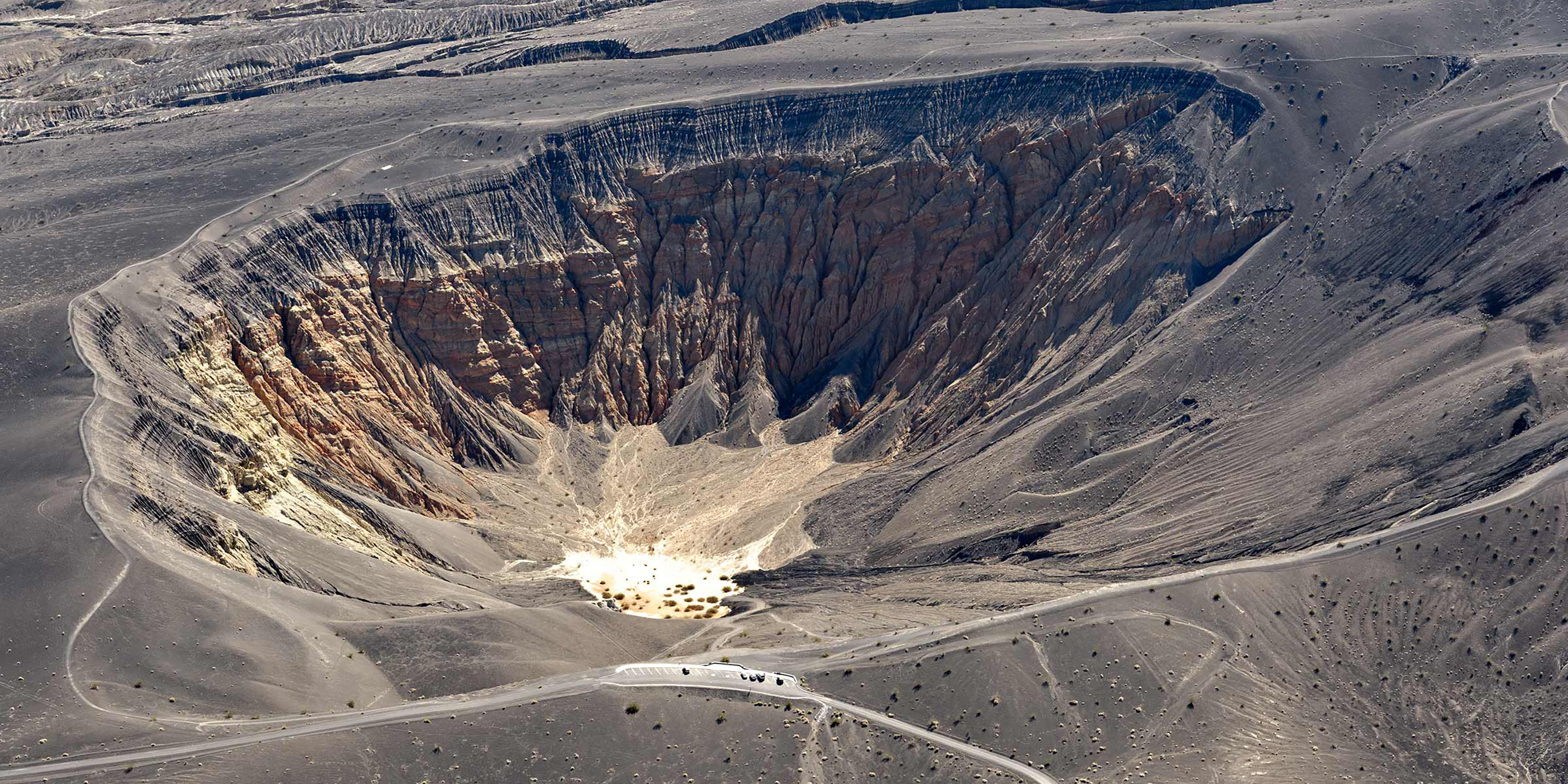 The photograph presents a vast, barren crater with steep, rocky sides and a pale, sandy bottom. It places emphasis on the crater's geological features, color stratifications, and visible man-made structures at its base.