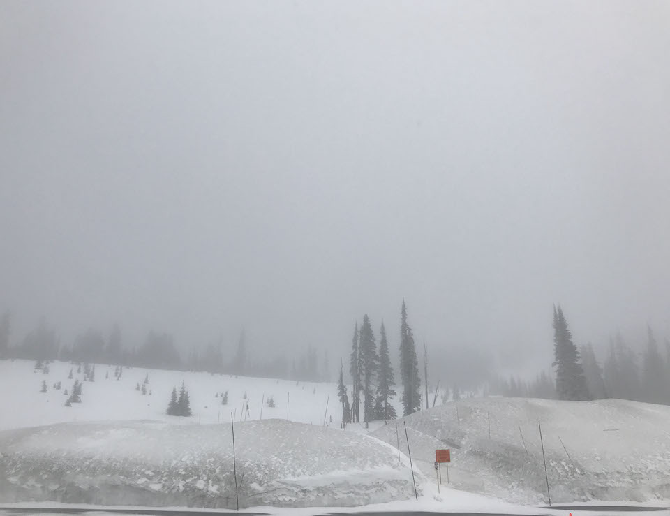 Profundos bancos de nieve rodean a una zona de estacionamiento, con los alrededores completamente ocultos por la niebla y nubes bajas.