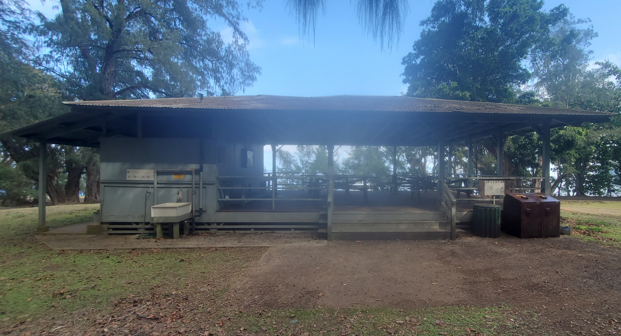 Outdoors; older pavilion in cut grass lawn with trees and blue sky behind.