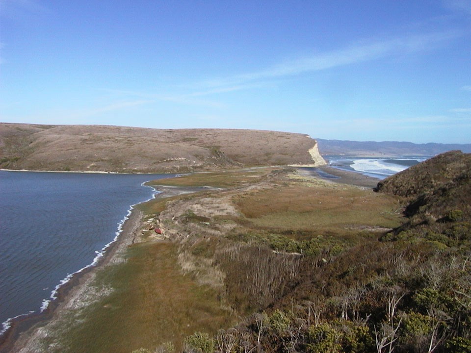 A pond on the left side of a grassy and sandy area with white-capped waves in a bay on the right.