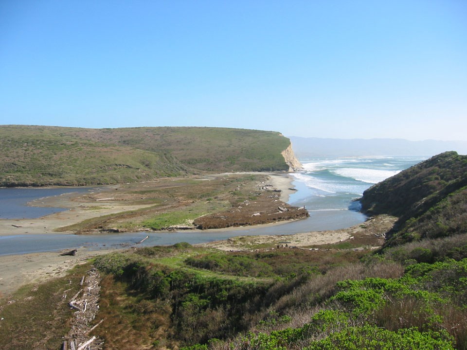 A pond on the left side of a grassy and sandy area with white-capped waves in a bay on the right.