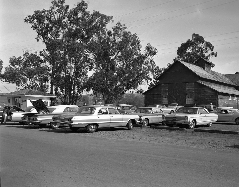 A black and white photo of 1960s automobiles parked to the right of a small building with an information sign and in front of an old barn.