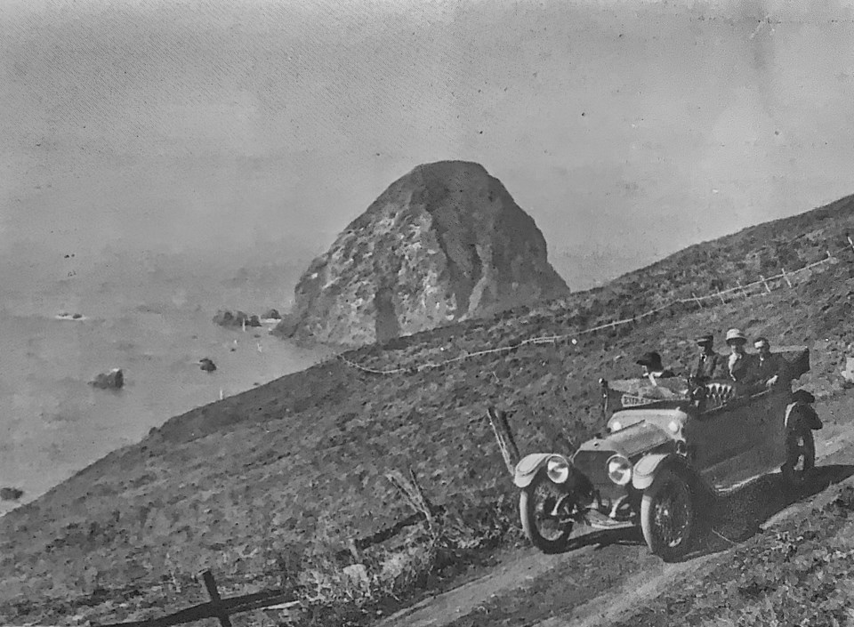 Old car on dirt road with large rock in ocean in background