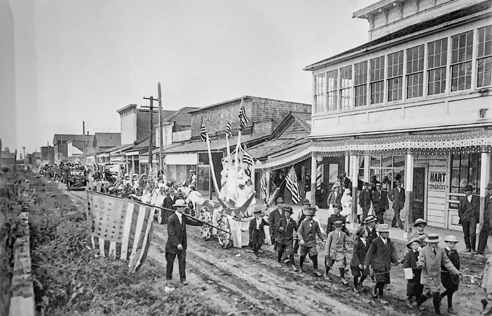 Parade with flags on dirt street in front of buildings