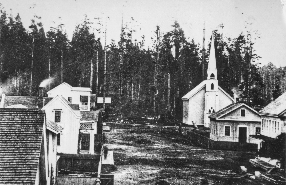 Church with steeple, dirt road, buildings and forest.