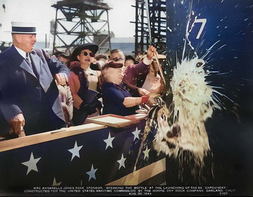 A group of men and women in dress clothes celebrate by breaking a champagne bottle across the ship.