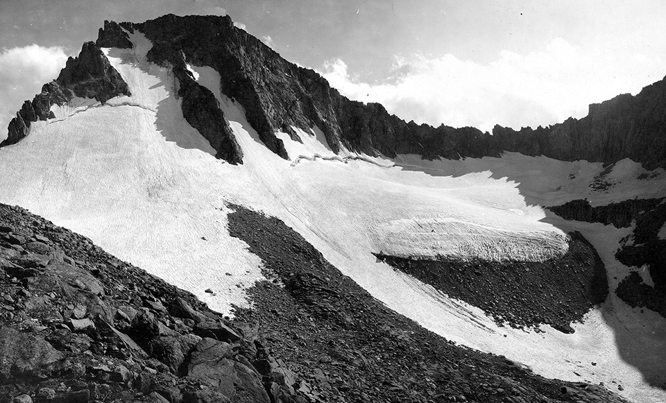 Rocky mountain slope mostly covered with a glacier that stretches out to both sides and has a chute stretching down toward lower righthand corner of photo.