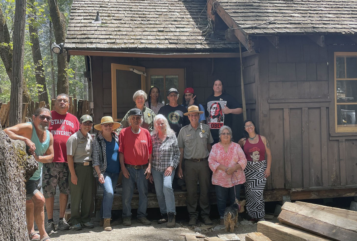 A black and white historical photo of a large group of people posing in front of a cabin in the New Village.