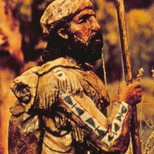 wood carving of bearded fur trapper in deerskin clothing, hat, and satchel standing in a field