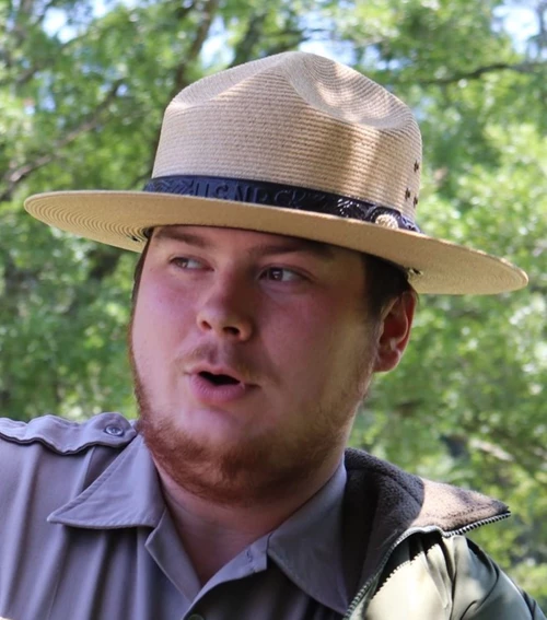 A ranger in a flat hat is speaking. There are trees in the background. 