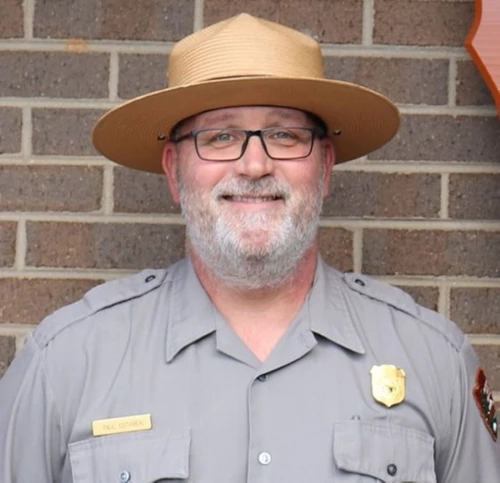 A ranger in a flat hat stands in front of a brick wall.