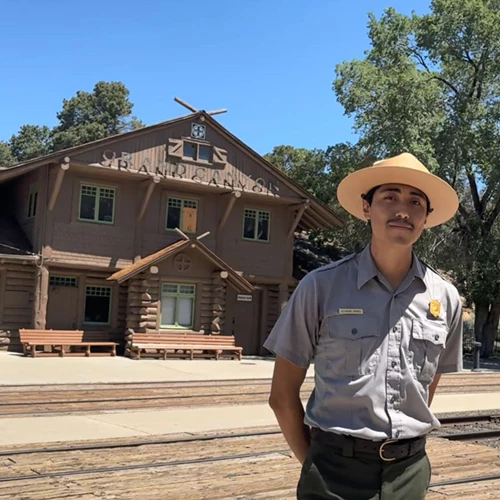 A ranger with a straw flat brimmed hat and uniform stands in front of a train near a train station.