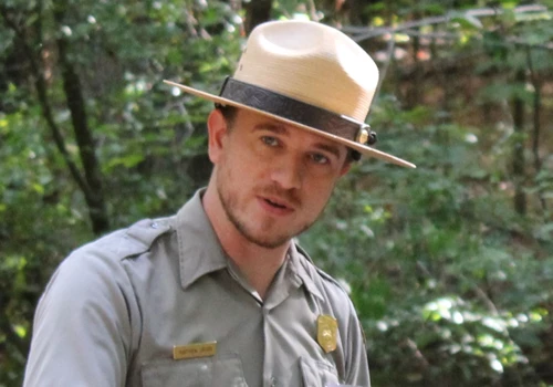 A park ranger in a flat hat speaks towards the camera. There are trees in the background. 