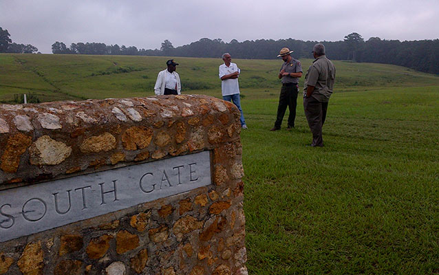 Park ranger and men stand in an open field area behind a stone monument labeled