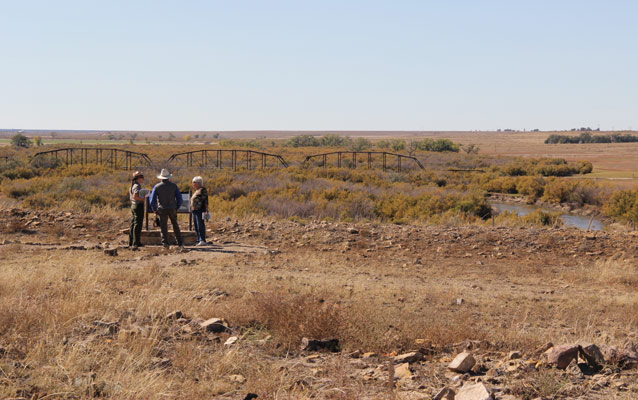 Three people stand in front of a wayside exhibit surrounded by fall grass, with a bridge and the Arkansas River.