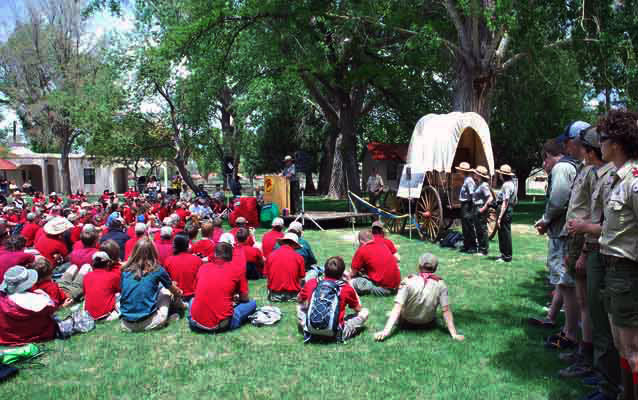 Boys in red shirts sit on a lawn, a speaker stands at a podium in a green uniform and straw hat.