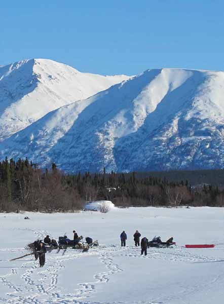 half a dozen people on a snowy field near a forest and steep, snowy mountains